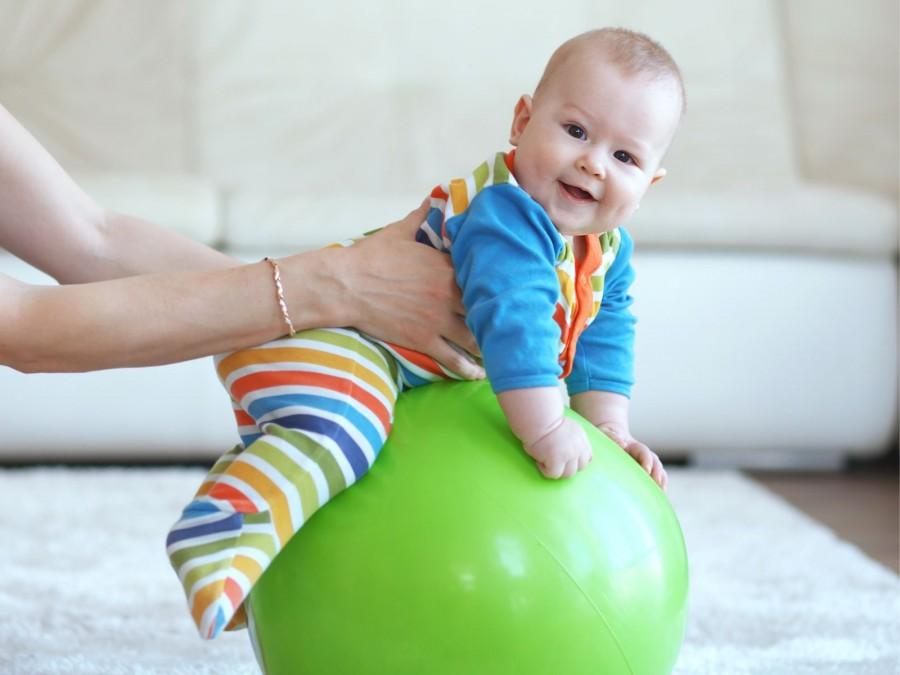 Ein lachendes Baby in einem bunten Strampelanzug sitzt auf einem grünem Gymnastikball und wird von dessen Mutter festgehalten.