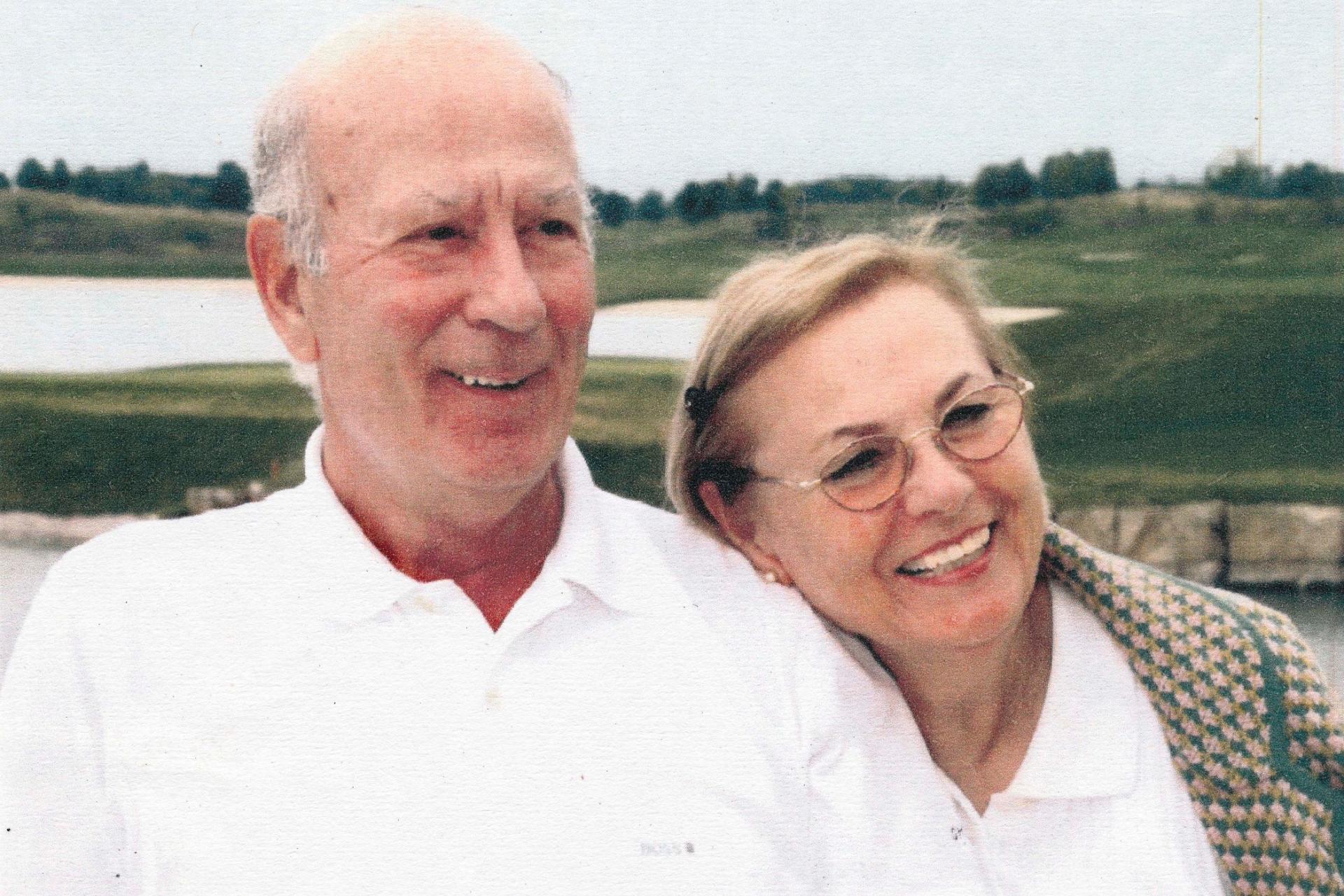Auf dem Foto ist ein älteres, lachendes Ehepaar abgebildet. Der Herr hat eine Halbglatze; das Haar ist bereits ergraut. Er trägt ein weißes Polo-Shirt. Die Frau lehnt ihren Kopf an seine Schulter. Sie hat mittelblonde Haare. Sie trägt eine Brille, eine weiße Bluse und einen karierten Blazer.