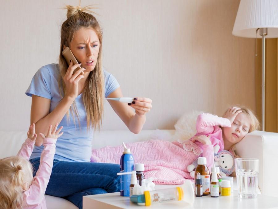 Eine Mutter telefoniert mit dem Notdienst und hält dabei ein Fieberthermometer in der Hand während ihre Tochter krank auf dem Sofa liegt. Die zweiter kleine Tochter steht neben der Mutter mit ausgestreckten Armen. Auf dem Wohnzimmertisch liegen verschiedene Medikamente.