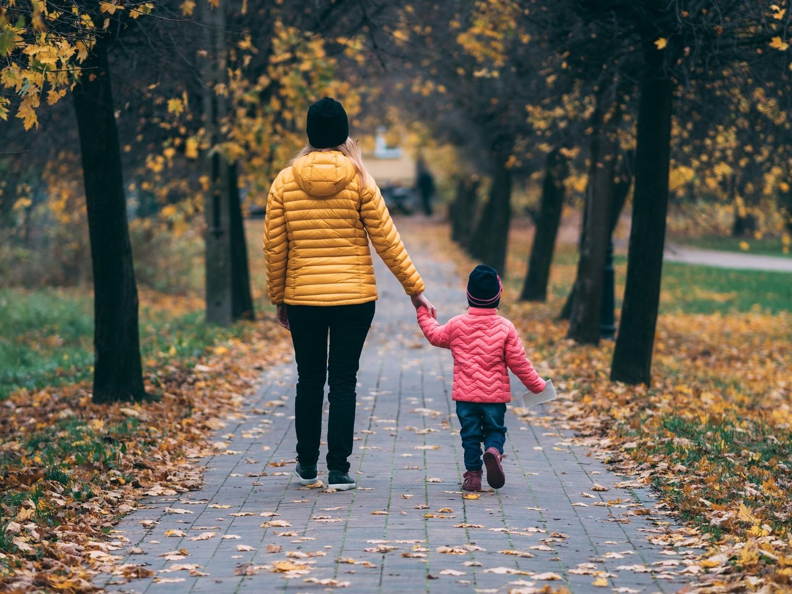 Frau mit kleiner Tochter an der Hand beim Spaziergang im Park, Laub liegt auf dem Weg.