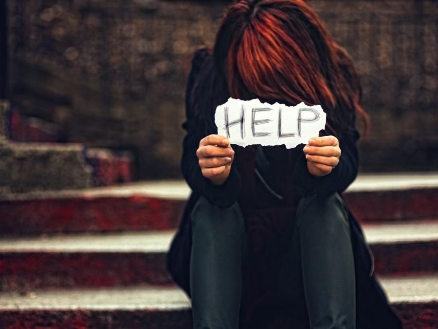 Eine Frau in schwarz gekleidet sitzt auf einer Treppe und hält einen Zettel mit der Aufschrift help in der Hand.