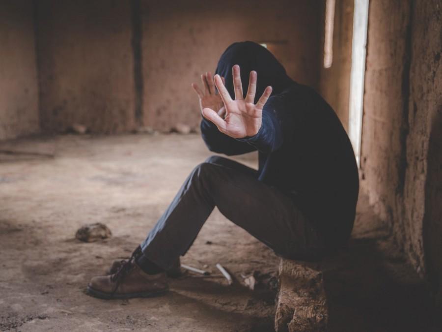 Ein Jugendlicher in einem schwarzen Hoodie mit Kapuze über dem Kopf sitzt in einem verlassenen Gebäude und zeigt eine Abwehrhaltung mit seinen Händen.