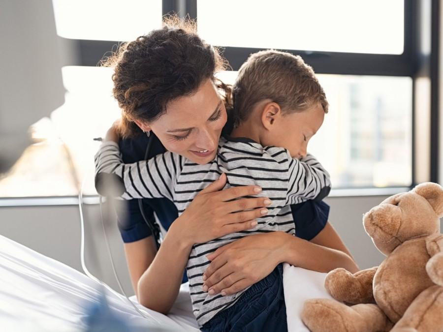 Eine Mutter umarmt ihren Sohn im Krankenhausbett. Rechts neben dem Kind sitzt ein brauner Teddy.