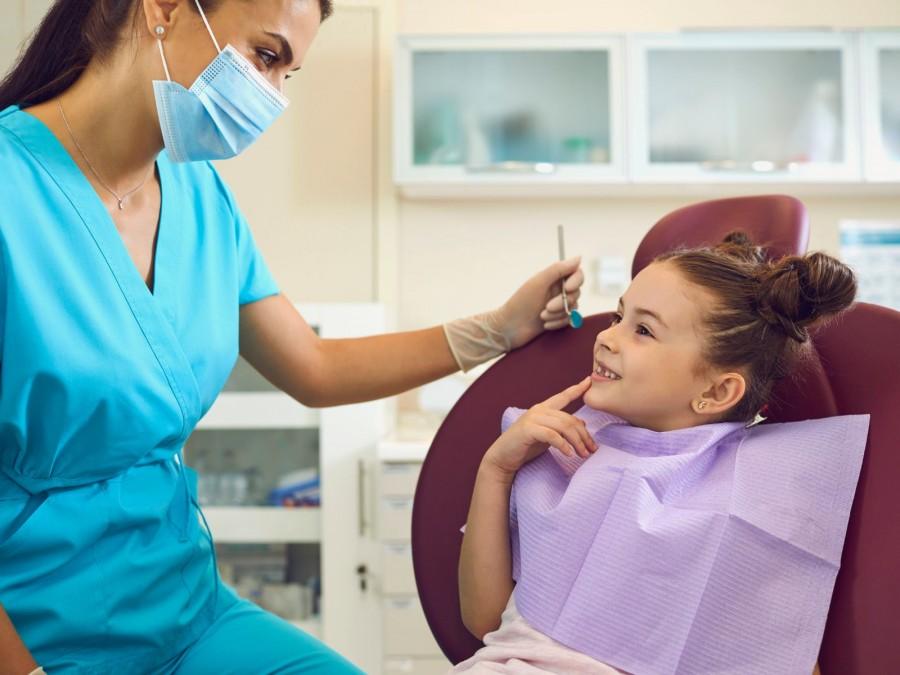 Ein junges Mädchen mit braunen Haaren und zwei Zöpfen sitzt lächelnd in einem Behandlungsstuhl in einer Praxis für Zahnmedizin. Gegenüber sitzt eine Zahnärztin in einer blauen Praxisbekleidung und hält in der linken Hand einen Mundspiegel.