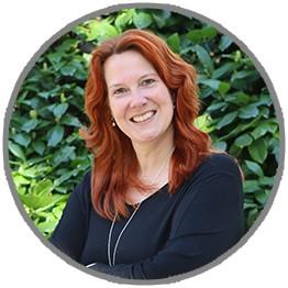 Frau mit roten, welligen Haaren trägt ein schwarzes Oberteil und silberne Halskette, vor grüner Hecke.