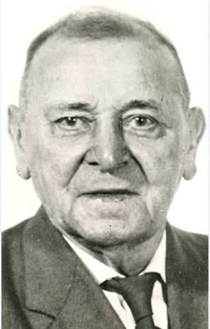 Auf dem Foto ist ein älterer Herr mit sehr kurzen Haaren zu sehen. Gekleidet ist er mit einer grauen Anzugjacke, einem weißen Hemd und einer schwarzen Krawatte.