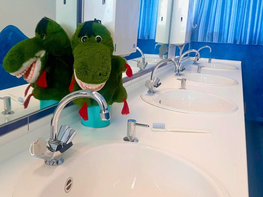 Ein Drache mit einem Gebiss sitzt als Stofftier auf einem Waschbecken im Mundhygienezentrum. Neben ihm liegt eine Zahnbürste.