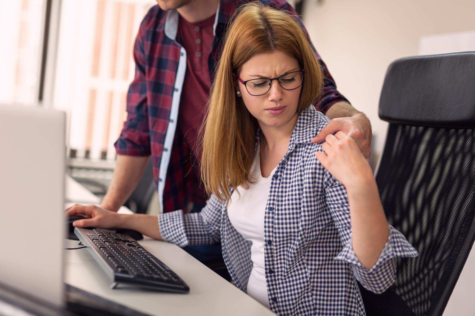Bild zeigt eine junge Frau am PC-Arbeitsplatz, ein Kollege stützt sich auf ihrem Schreibtisch ab und hat einen Arm um sie gelegt, den sie verägert wegschiebt.