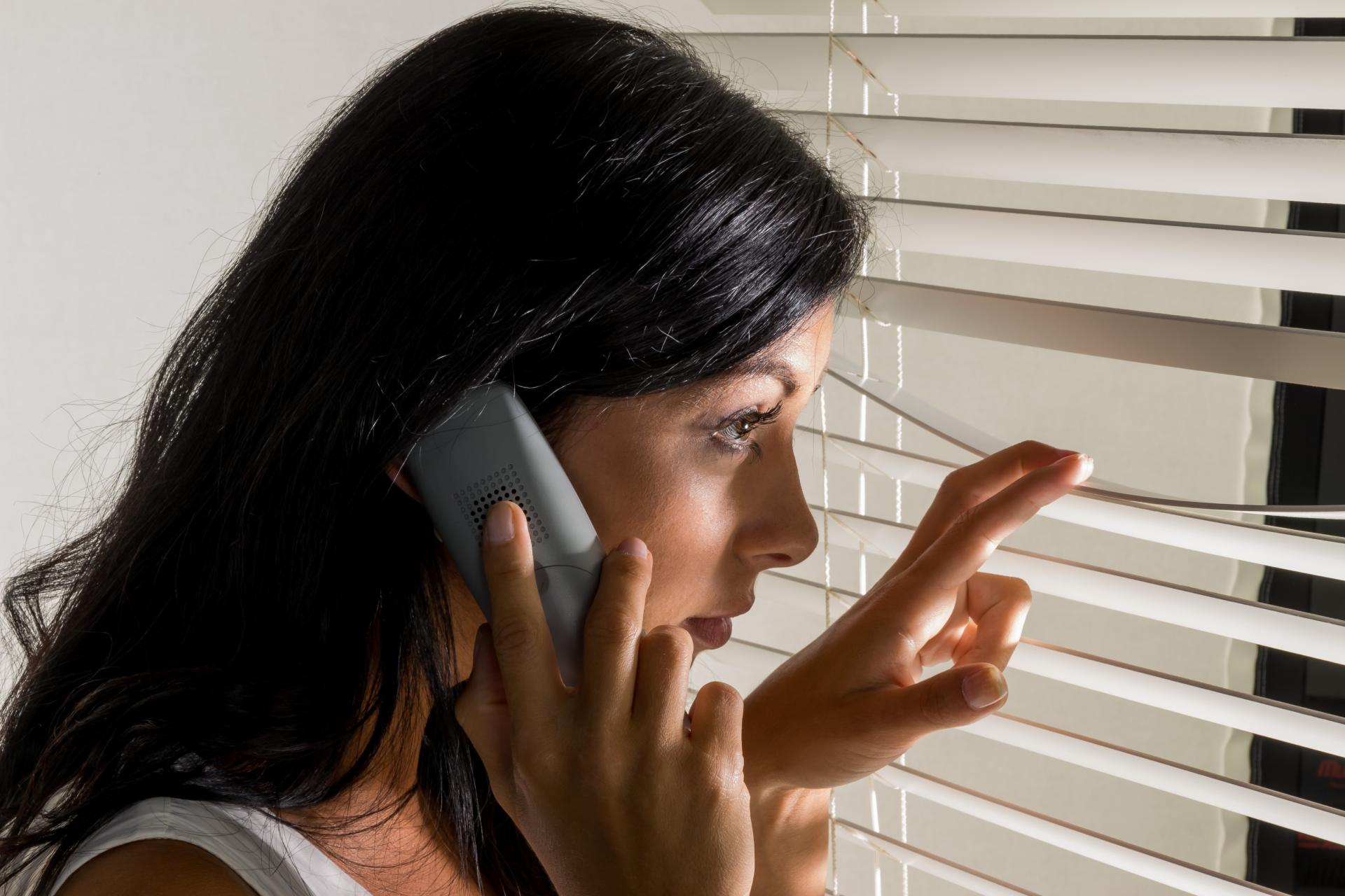 Junge Frau mit Telefon am Ohr schaut besorgt durch die Rollos an ihrem Fenster. Stalking-Handlungen beeinträchtigen den Tagesablauf und die Lebensqualität der Opfer erheblich.