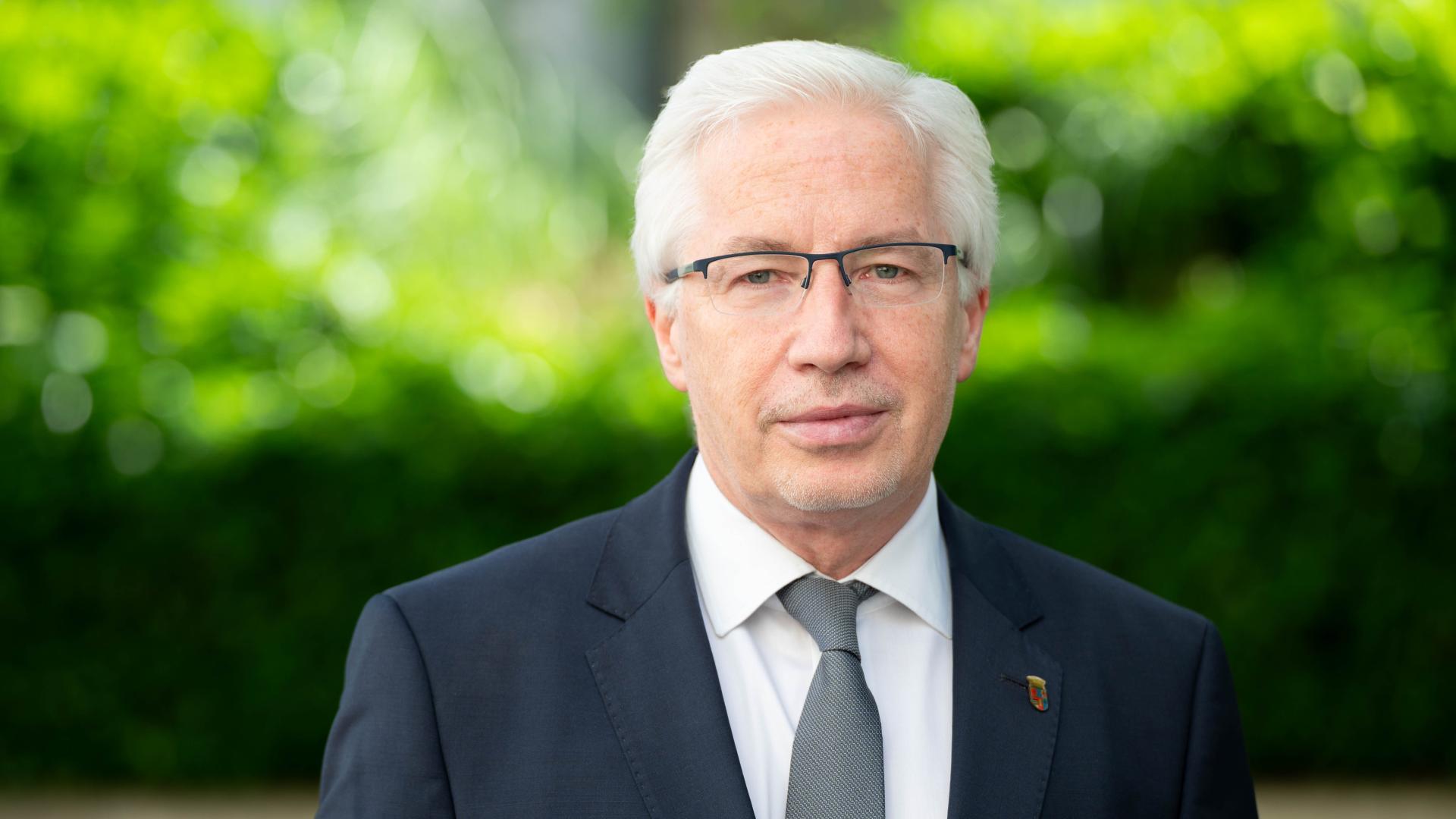 Mann mit weißem Haar trägt dunklen Anzug, weißes Hemd und graue Krawatte, steht vor grünem Hintergrund