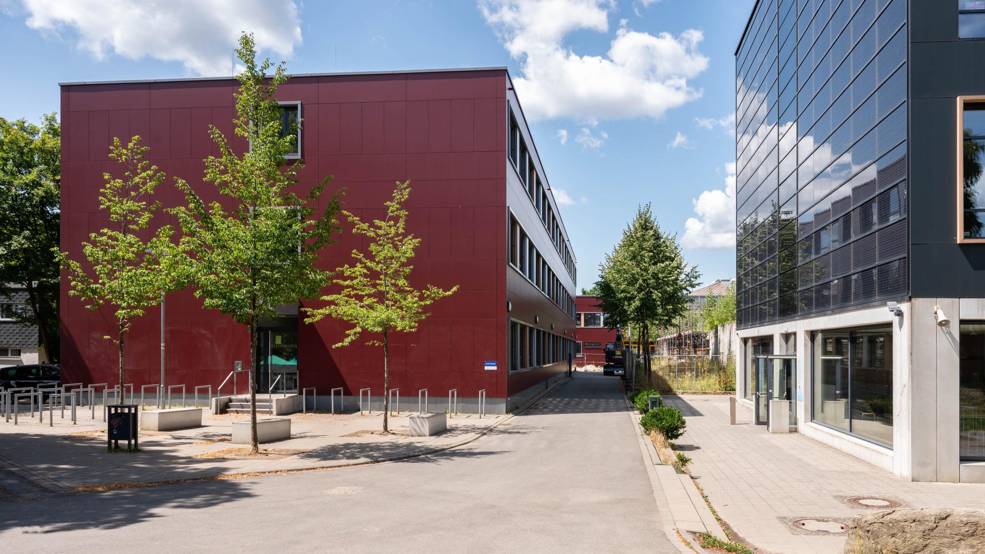 Schulgebäude mit roter Fassade links und modernes Gebäude mit Glasfassade rechts, dazwischen Gehweg und Bäume unter blauem Himmel