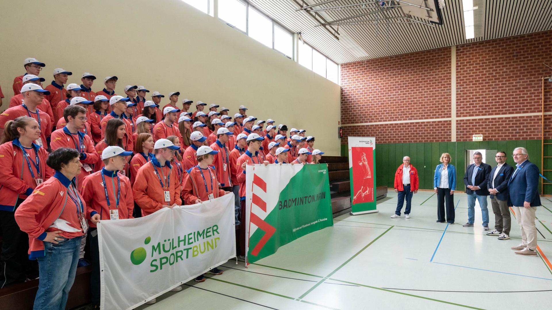 Große Gruppe junger Personen in roten Jacken und weißen Mützen steht in einer Sporthalle vor vier Erwachsenen, die sprechen. Banner mit Mülheimer Sportbund und Badminton sind sichtbar.