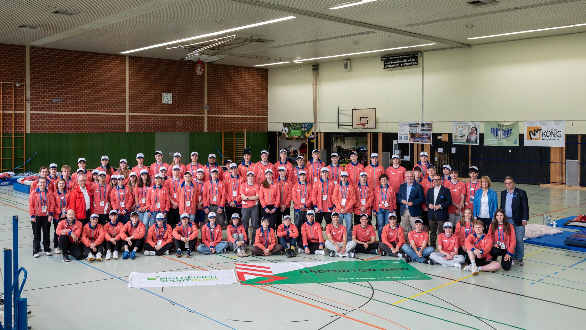 Große Gruppe von Jugendlichen und Erwachsenen in Sportkleidung und roten Jacken in der Sporthalle Von-der-Tann-Straße, Gruppenfoto beim FISU Badminton Youth Camp
