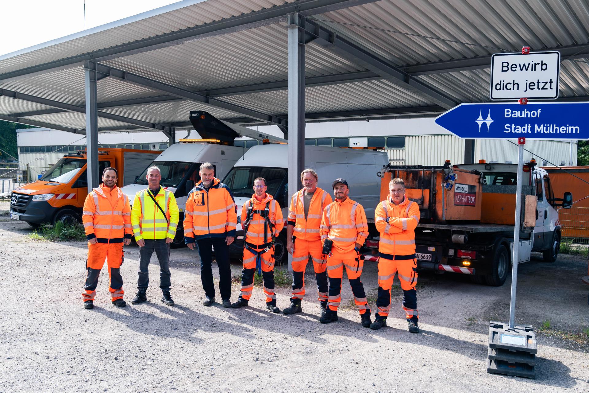 Mitarbeitende des Bauhofs in oranger und gelber Arbeitskleidung stehen vor einem Unterstand mit ihren Fahrzeugen. Neben ihnen steht eine blaues Richtungsschild mit der Aufschrift Bauhof Stadt Mülheim. Darüber ist ein weißes Schild mit dem Satz "Bewirb dich jetzt" montiert.