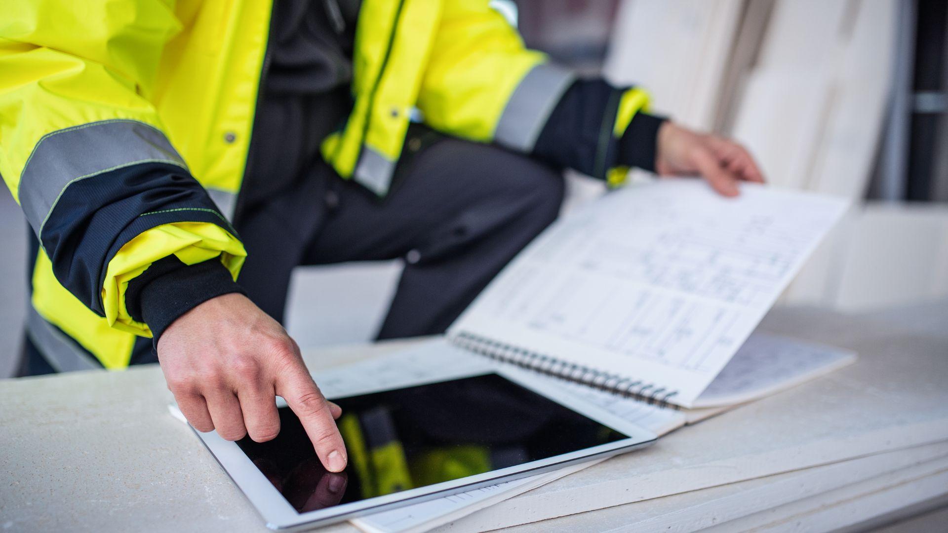 Ein Arbeiter in gelber Arbeitsmontur gleicht Notizen in einem Heft mit seinem Tablet ab.