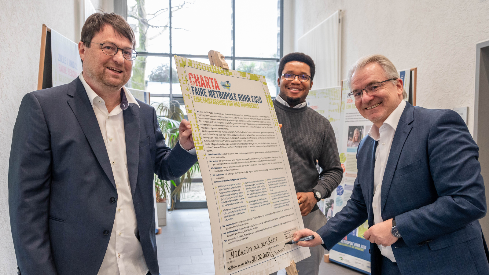 Drei Personen in Anzügen stehen in einem hellen Raum, einer hält ein großes Plakat mit der Überschrift Charta Faire Metropole Ruhr 2020 und unterschreibt es