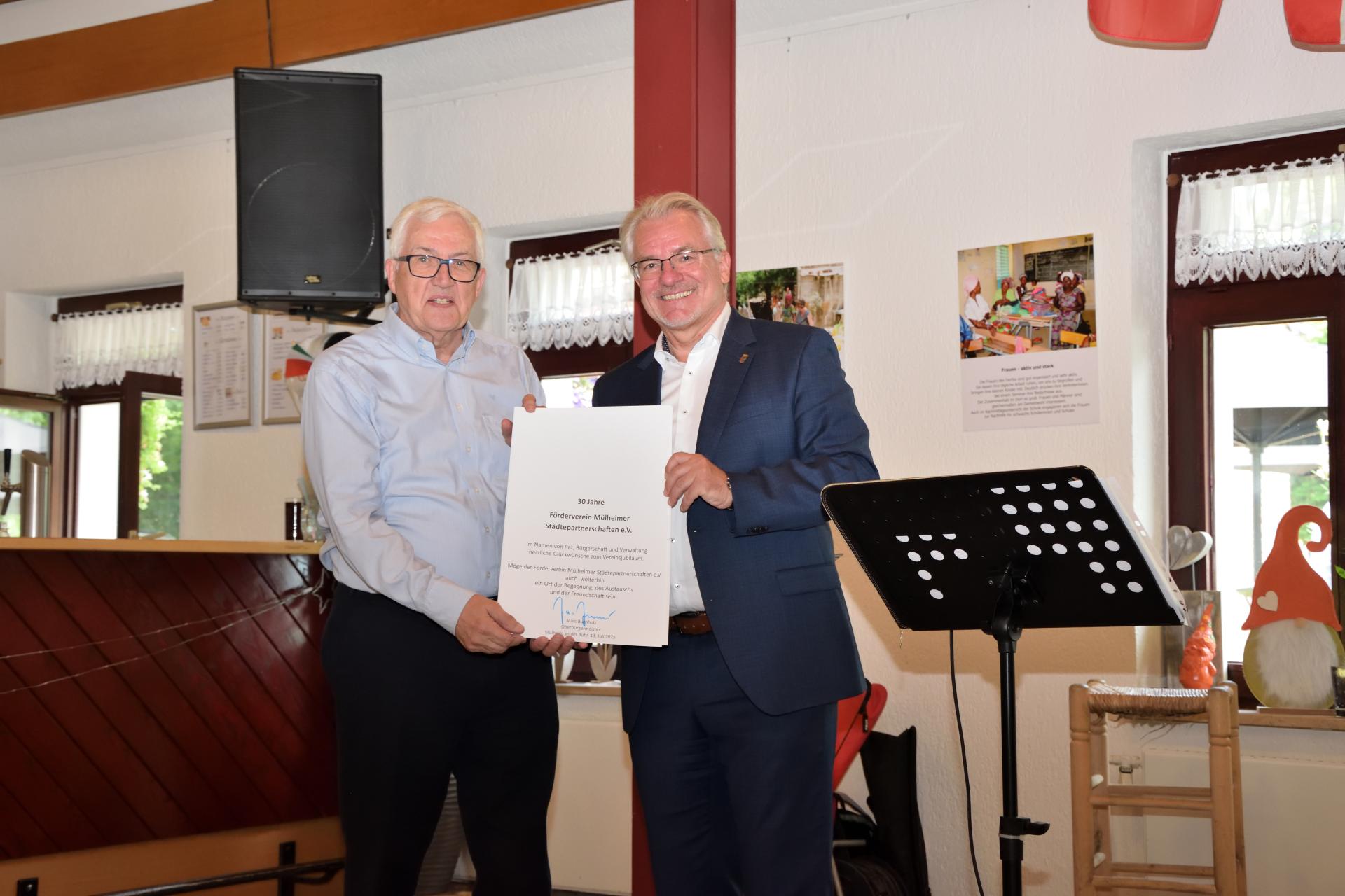 Oberbürgermeister Marc Buchholz (rechts) überreicht dem Vorsitzenden des Fördervereins Mülheimer Städtepartnerschaften e. V., Dr. Gerhard Ribbrock (links), eine Urkunde anlässlich des 30-jährigen Jubiläums des Vereins.
