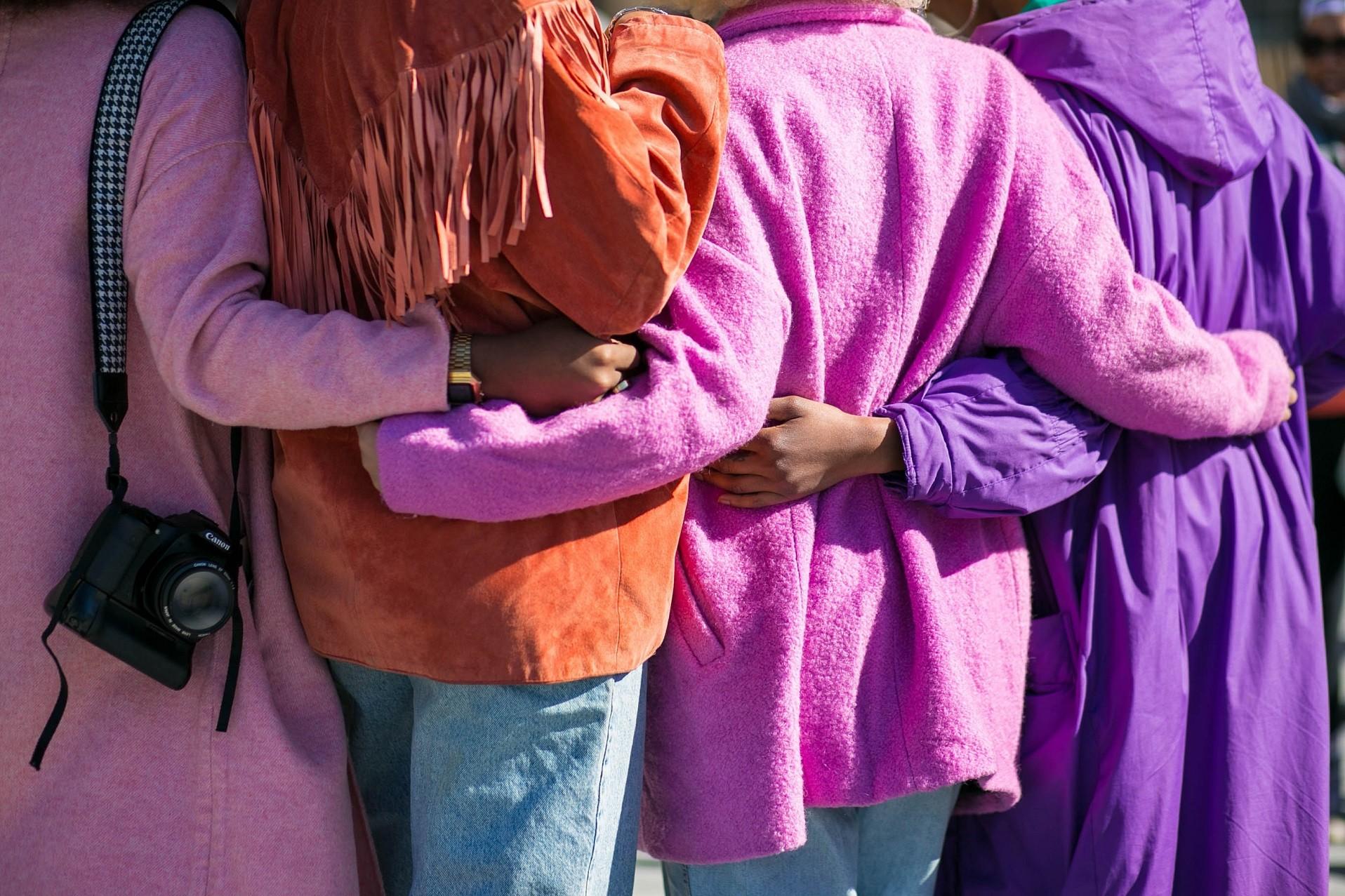 Von hinten fotografiert: Vier Frauen in knallbunten Mänteln halten sich in den Armen, Arm in Arm. Symbol für Gleichstellungsstelle, Zusammenhalt