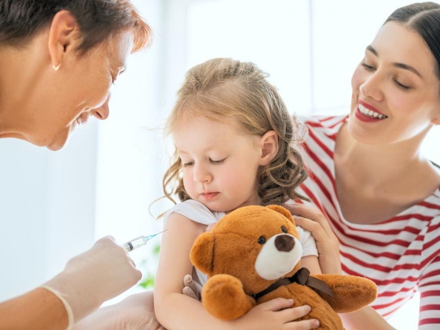 Eine Mutter hat Ihre Tochter auf dem Schoß. Das Mädchen hält einen Teddybären im Arm während eine Ärztin das Kind impft.