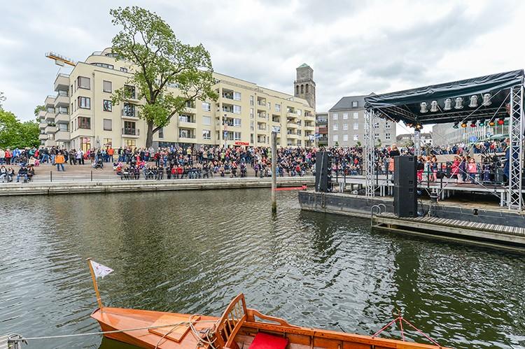 Blick vom Wasser aus in den Ruhrbania-Hafen anlässlich der offiziellen Eröffnung im Jahr 2014. Eine Bühne ist aufgebaut, Menschen stehen am Ufer. Im Hintergrund Bebauung und der Rathausturm.