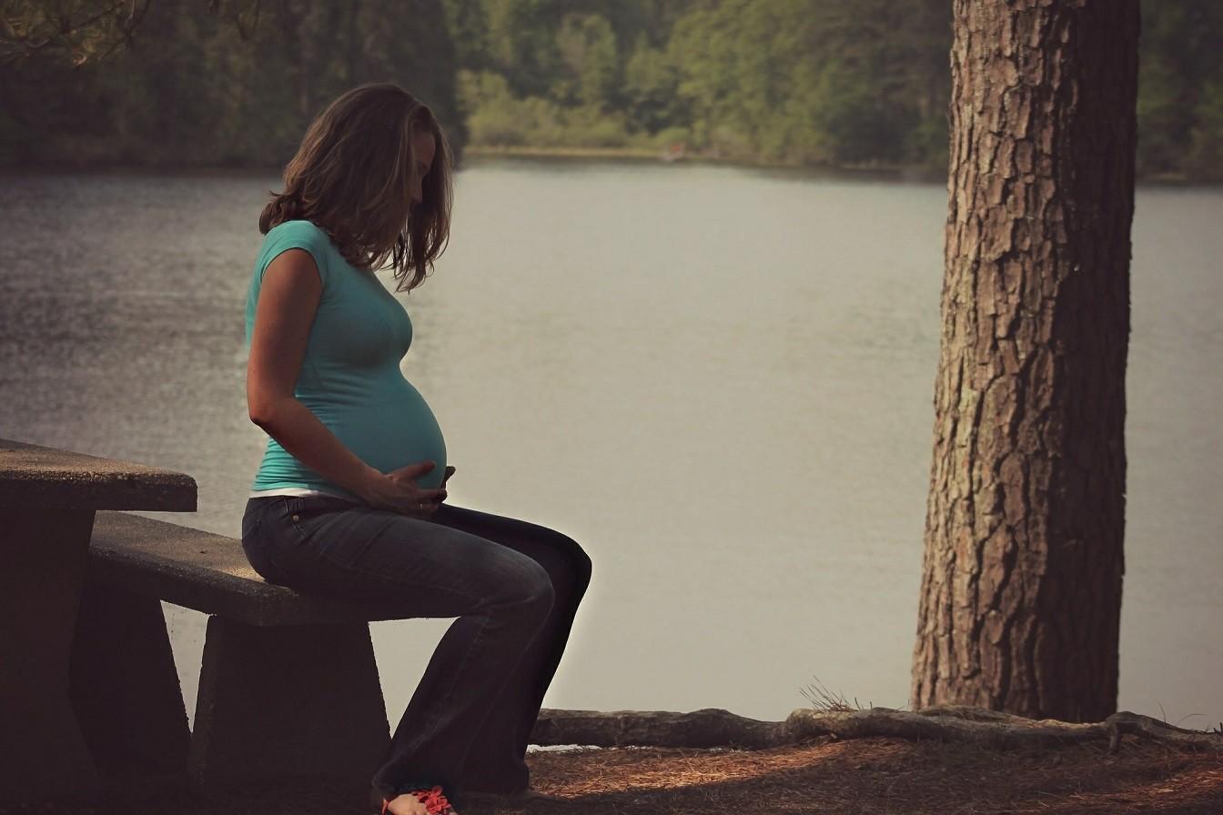Eine schwangere Frau sitzt auf einer Holzbank an einem See.