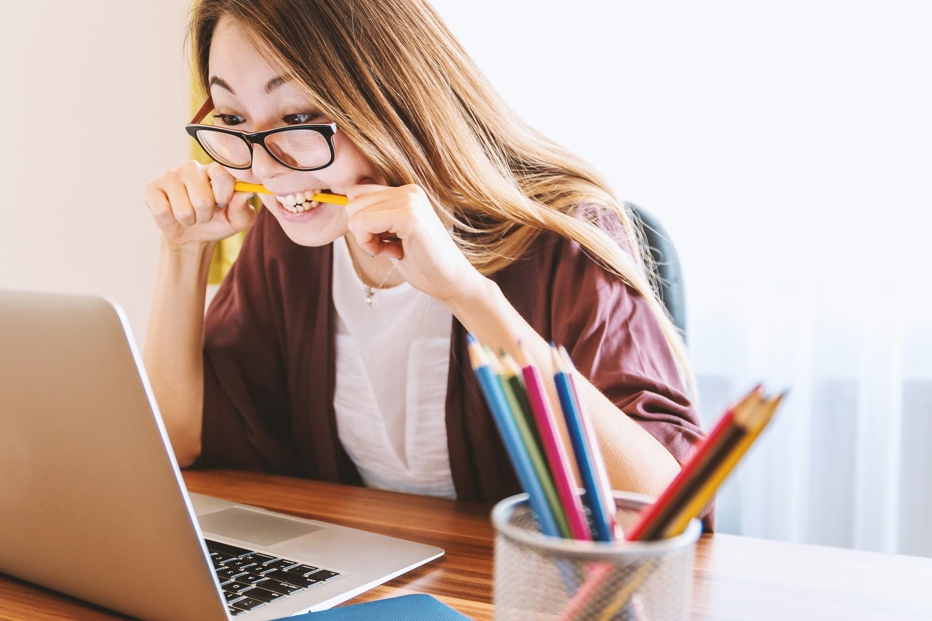 Junge Studierende sitzt vor aufgeklapptem Laptop, schaut auf den Bildschirm und beißt auf einen Stift