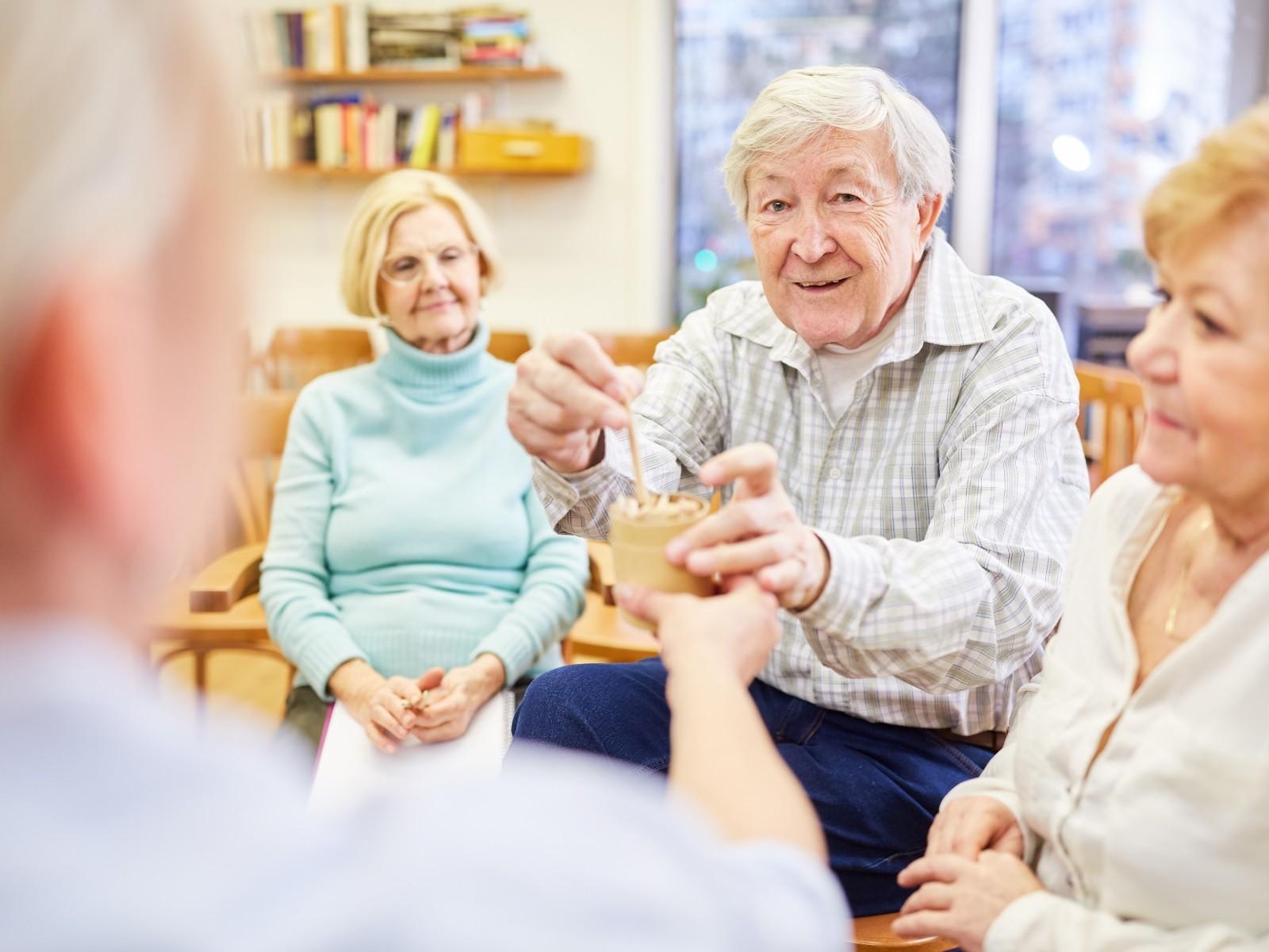 Auf dem Foto ist eine Gruppe von Seniorinnen und Senioren in einem Lehrraum zu sehen. Ein älterer Mann nimmt dabei von seinem Gegenüber (nur als Hinteransicht) ein Getränk entgegen.