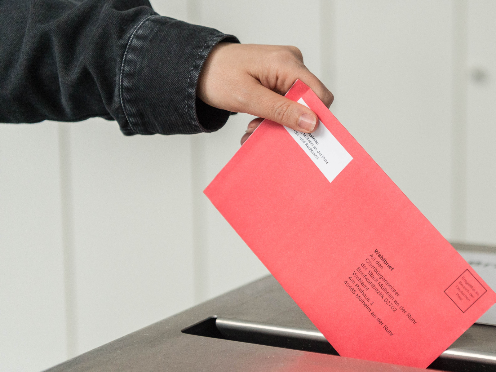 Eine Hand wirft den versiegelten roten Wahlumschlag mit dem Stimmzettel in den Schlitz der Wahlurne.