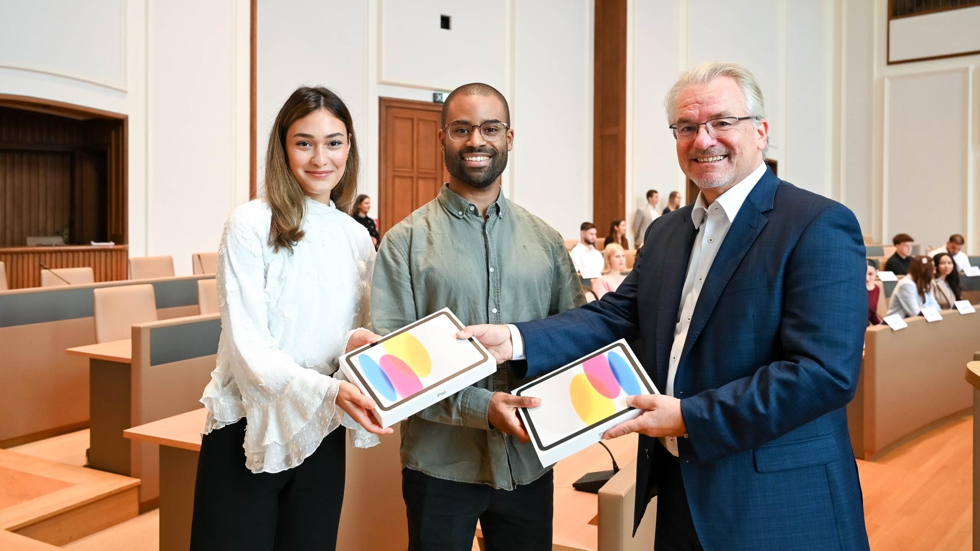 Oberbürgermeister Marc Buchholz übergibt zwei Tablets an zwei Auszubildende.