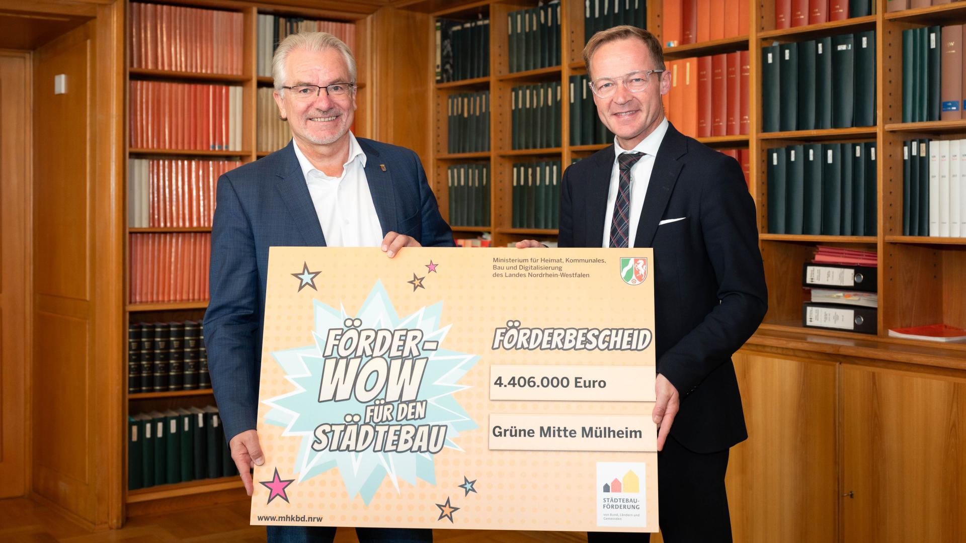Oberbürgermeister Marc Buchholz und Staatssekretär Daniel Sieveke halten einen großen Förderbescheid mit der Aufschrift 4.406.000 Euro, Grüne Mitte Mülheim in der Rathausbibliothek.