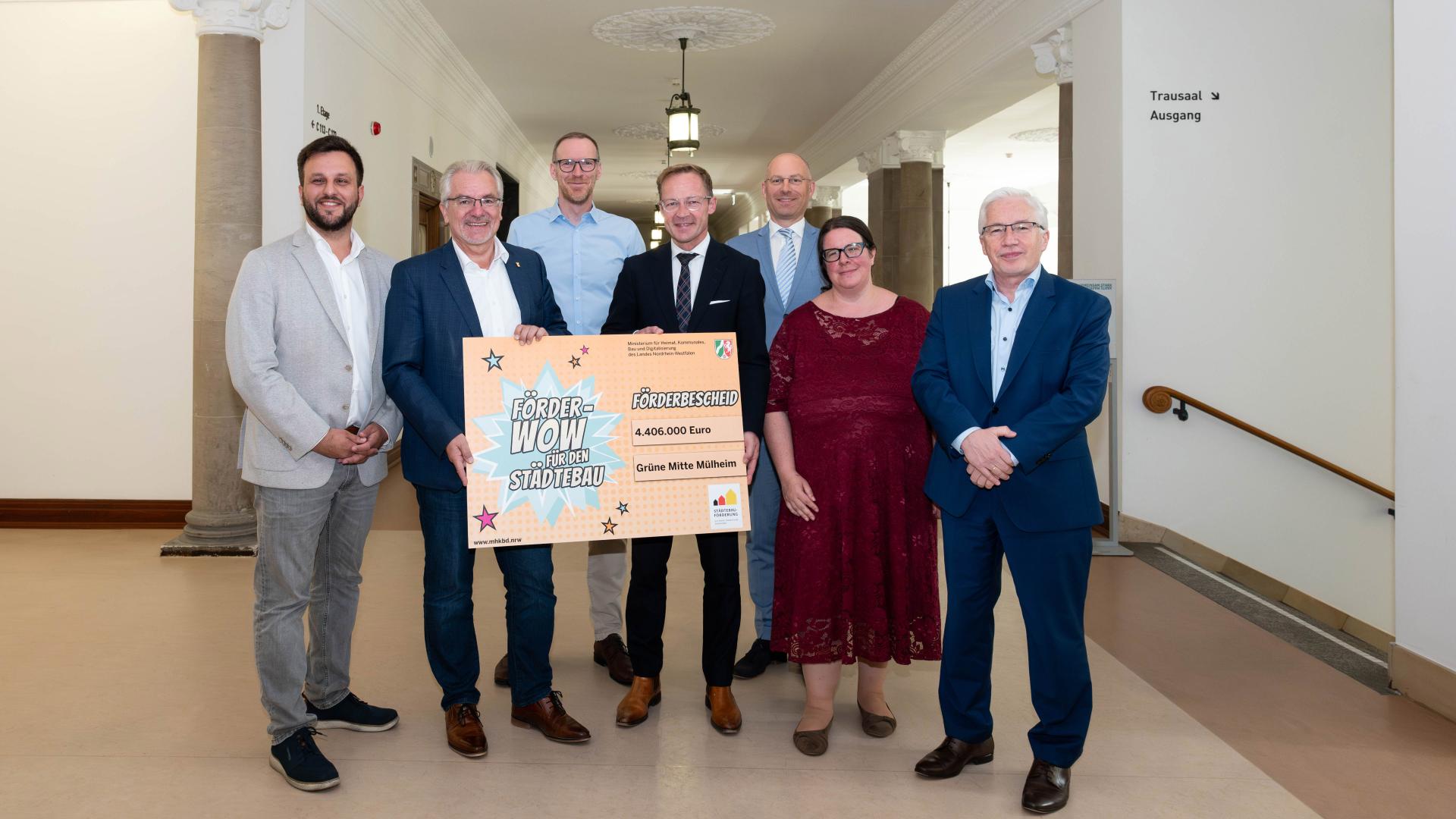 Sieben Personen stehen in einem Flur, Oberbürgermeister Marc Buchholz und Staatssekretär Daniel Sieveke halten einen großen Förderbescheid mit der Aufschrift FÖRDER WOW STÄDTEBAU und 4.406.000 Euro.
