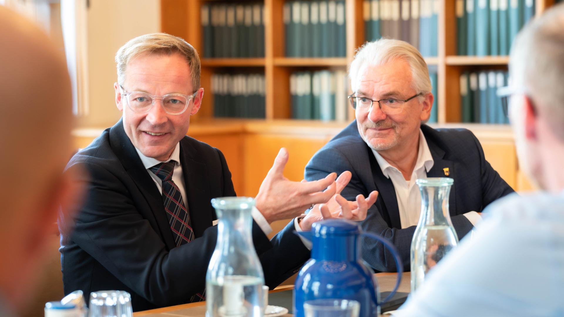 Staatssekretär Daniel Sieveke im Gespräch mit Oberbürgermeister Marc Buchholz, an einem Tisch sitzend mit Karaffen und Gläsern, Bücherregal im Hintergrund