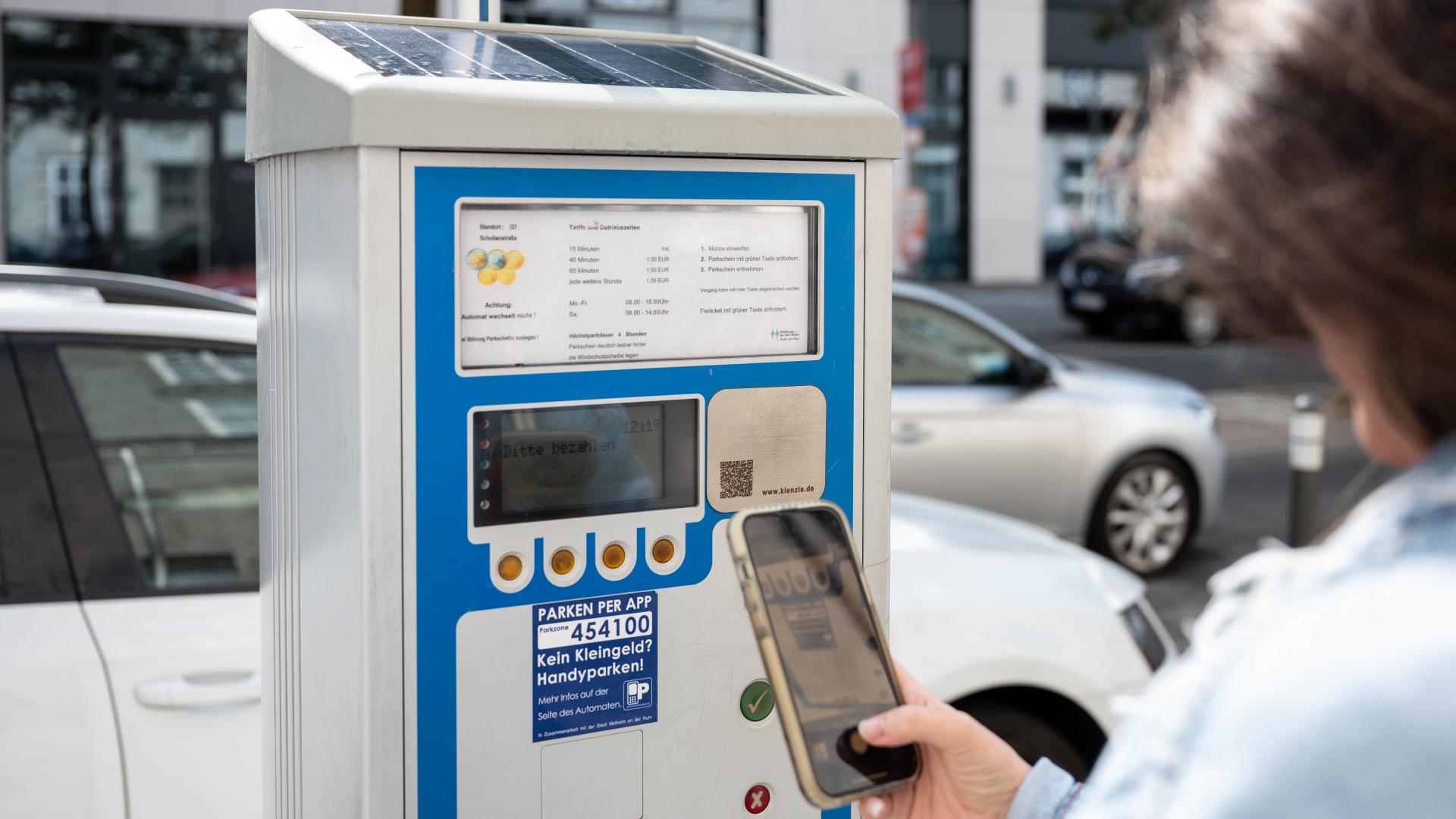Parkscheinautomat mit Solarzelle, Person hält Smartphone in der Hand, auf dem Display ist ein QR-Code sichtbar.