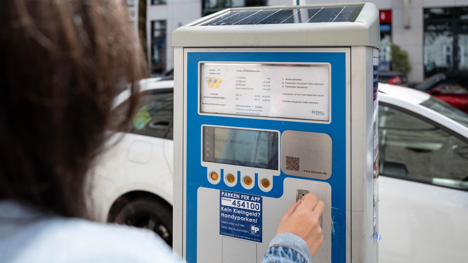 Parkscheinautomat mit blauer und grauer Oberfläche, Person tippt auf das Bedienfeld, QR-Code und Anweisungen sichtbar.