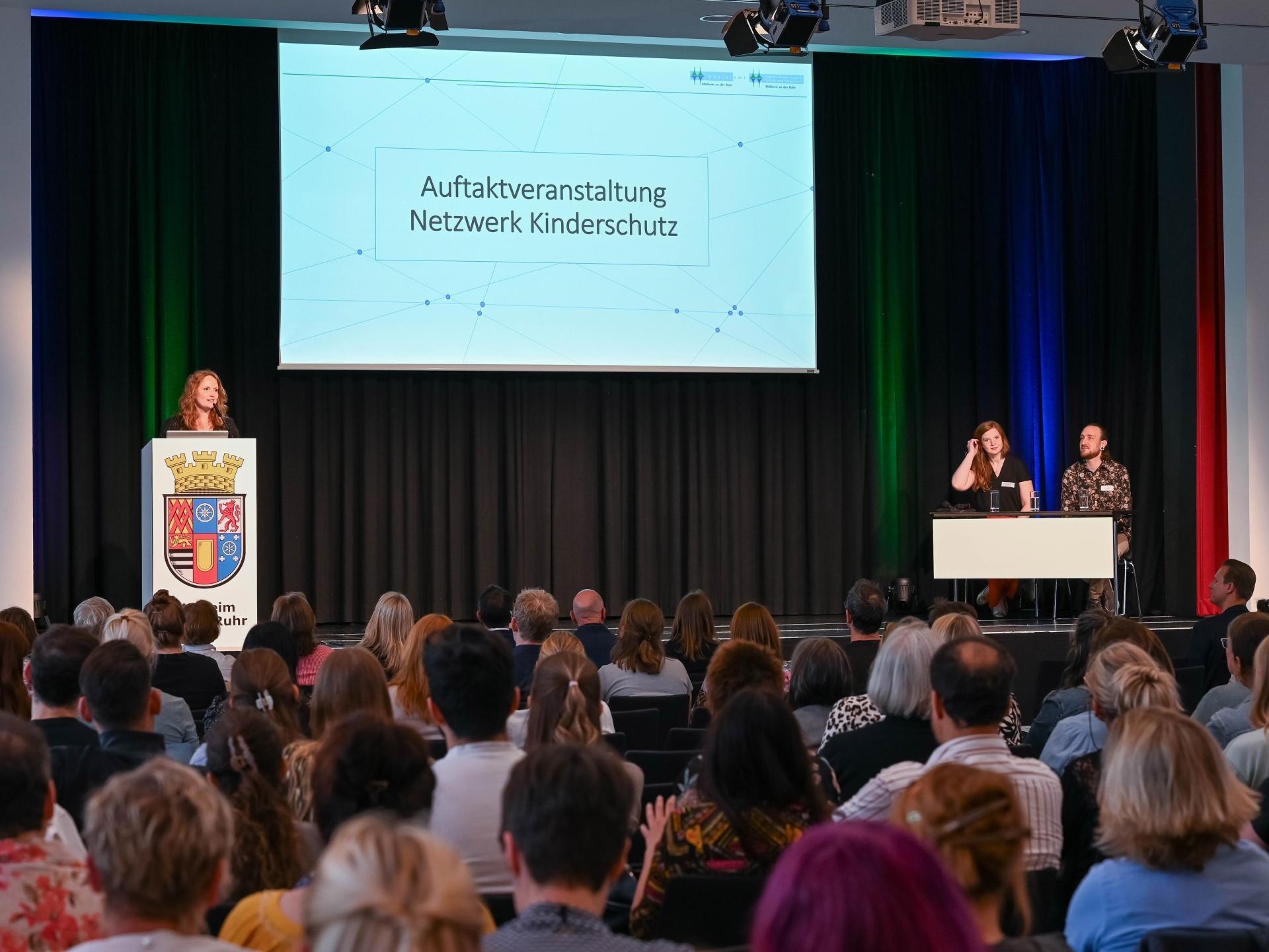 Auf der Bühne begrüßt Julia Brunner zusammen mit ihrem Team (Lara Vercoulen und Heiko Möller) die Teilnehmenden der Auftaktveranstaltung Netzwerk Kinderschutz