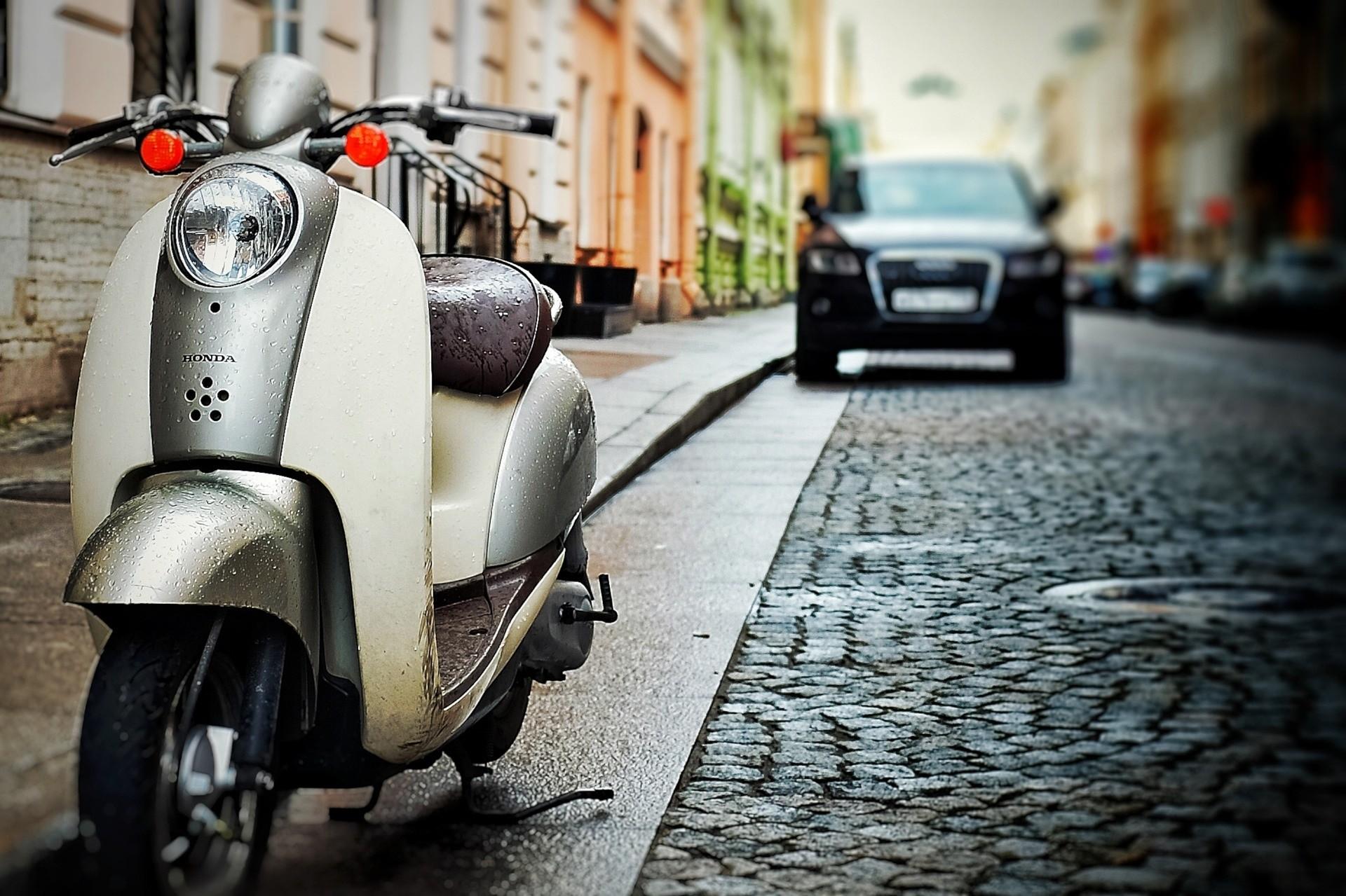 Das Foto zeigt einen Motoroller und ein Auto geparkt in einer Straße mit Kopfsteinpflaster umringt von Altbauten.