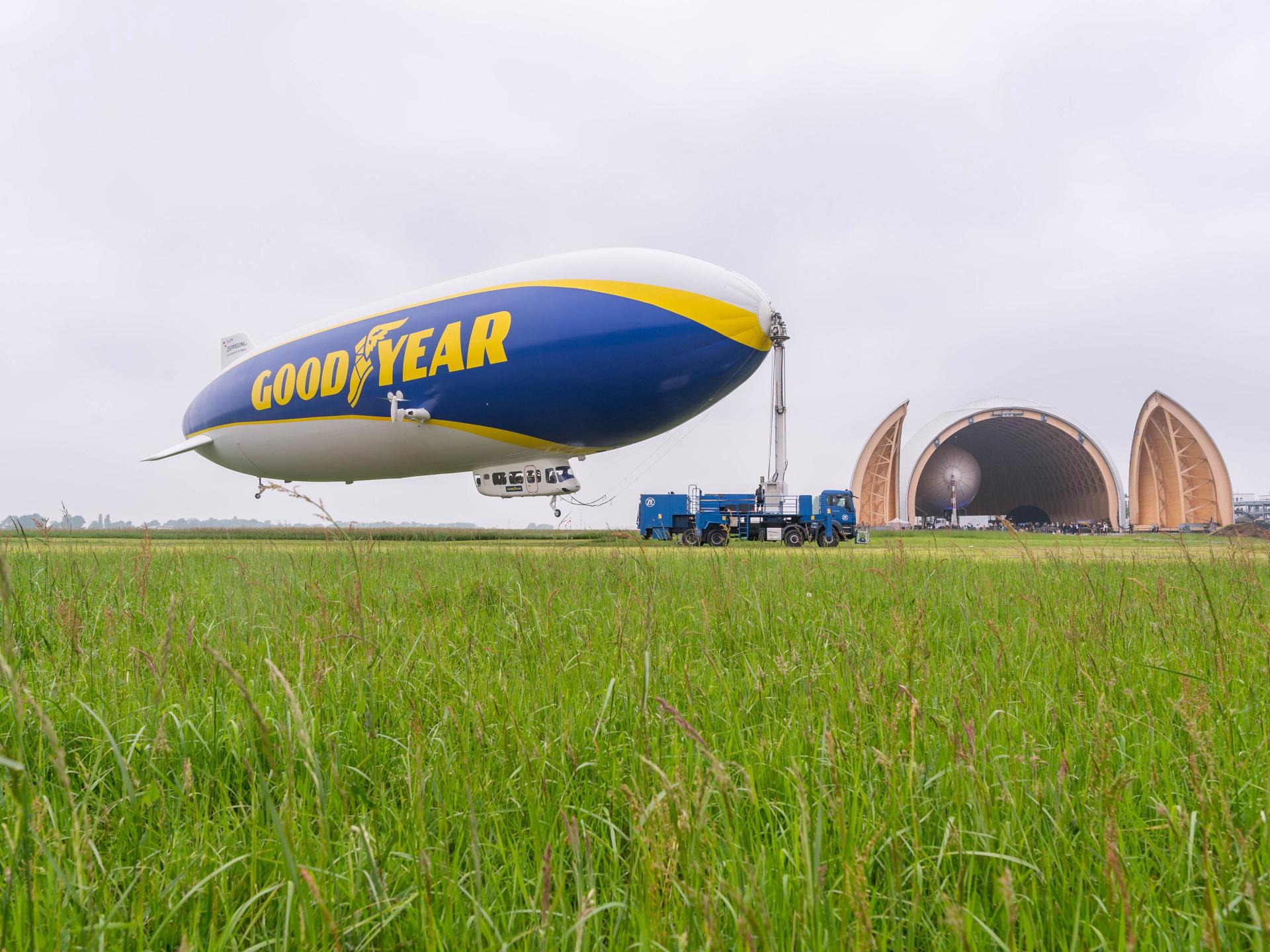 Blick über eine grüne Wiese am Flughafen Essen/Mülheim auf den schwebenden Zeppelin NT neben der Luftschiffhalle.