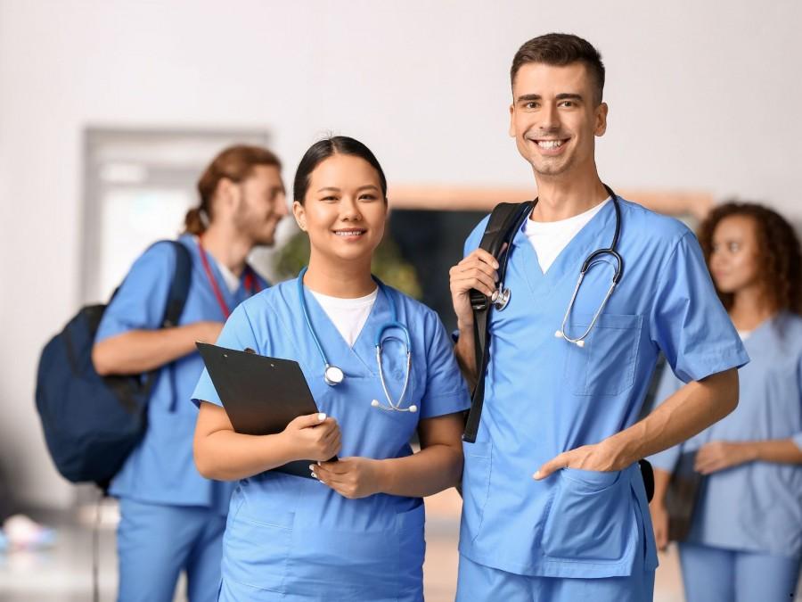 Eine Medizinstudentin und ein Medizinstudent stehend lächelnd in der Universität. Im Hintergrund befinden sich zwei weitere Student*innen.