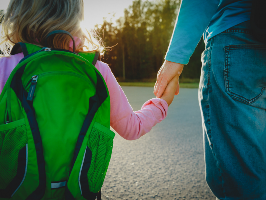 Bildausschnitt mit einem Elternteil, der sein kleines Kind mit grünem Rucksack an der Hand hält und aus dem Bild laufen. Tagespflegedienst, Kita, Kindergarten, Kinderbetreuung, Pflegevater, Pflegemutter, Kindertagespflege.
