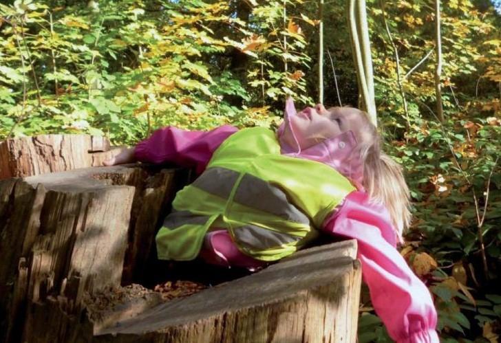 Kleines Mädchen in gelber Warnweste liegt zwischen Baumstümpfen, schaut hoch in den Himmel, in der Parkanlage Witthausbusch und genießt die Natur mit allen Sinnen