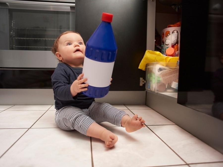 Kleinkind hat die Türen zum Putzmittelschrank geöffnet und hält eine Flasche mit giftigem, ätzendem Reinigungsmittel in den Händen.