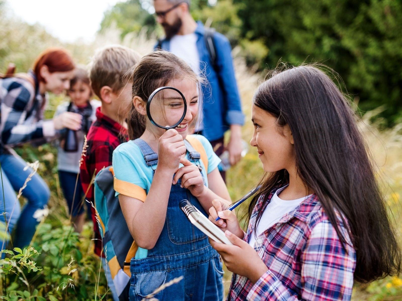 Ausflug mit Kindern und zwei Betreuungspersonen in der Natur. Im Vordergrund sind zwei Mädchen zu sehen, eines hält eine Lupe und das andere Mädchen einen Notizblock.