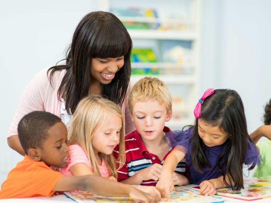 Weibliche Tagespflegeperson schaut mit vier Kindern ein Bilderbuch in ihrem Tagespflegenest an. Kindertagespflegeperson, Kita, Kindergarten, Kinderbetreuung, Pflegevater, Pflegemutter