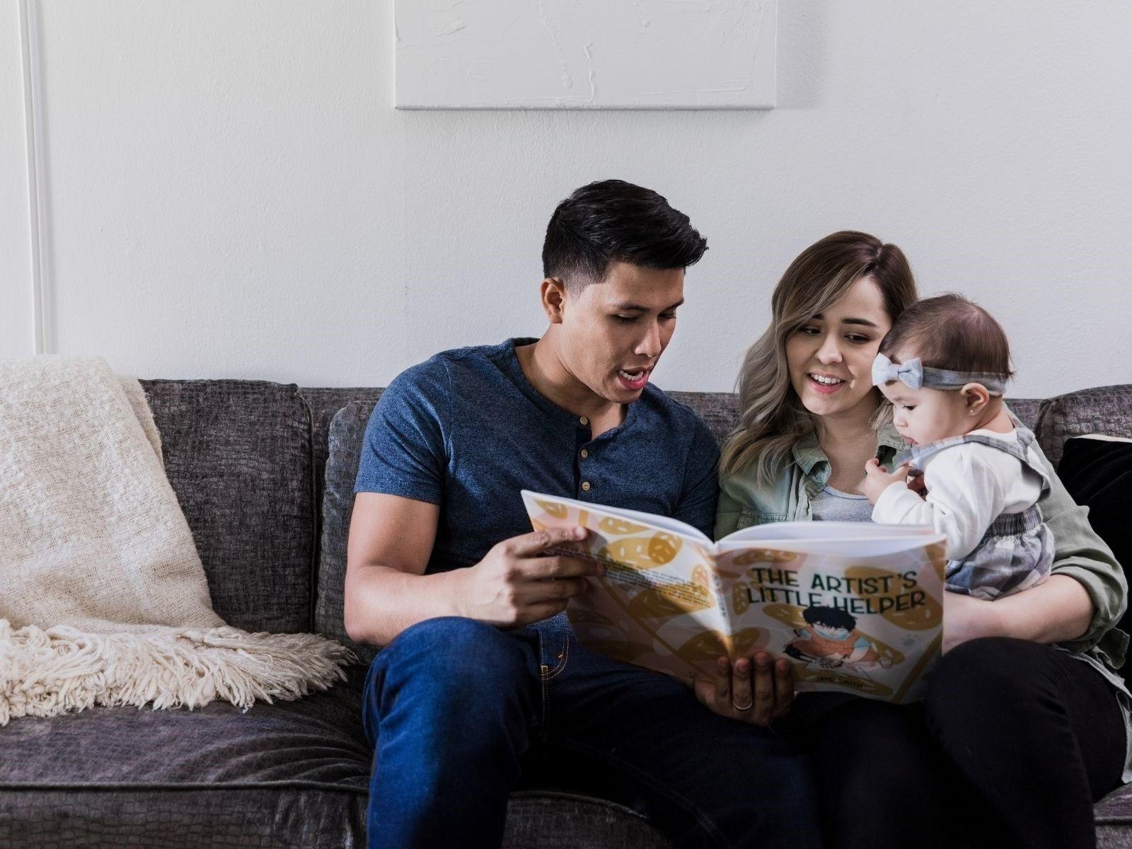 Junge Familie sitzt zusammen und die Eltern lesen ihrem Kleinkind aus einem Buch vor.
