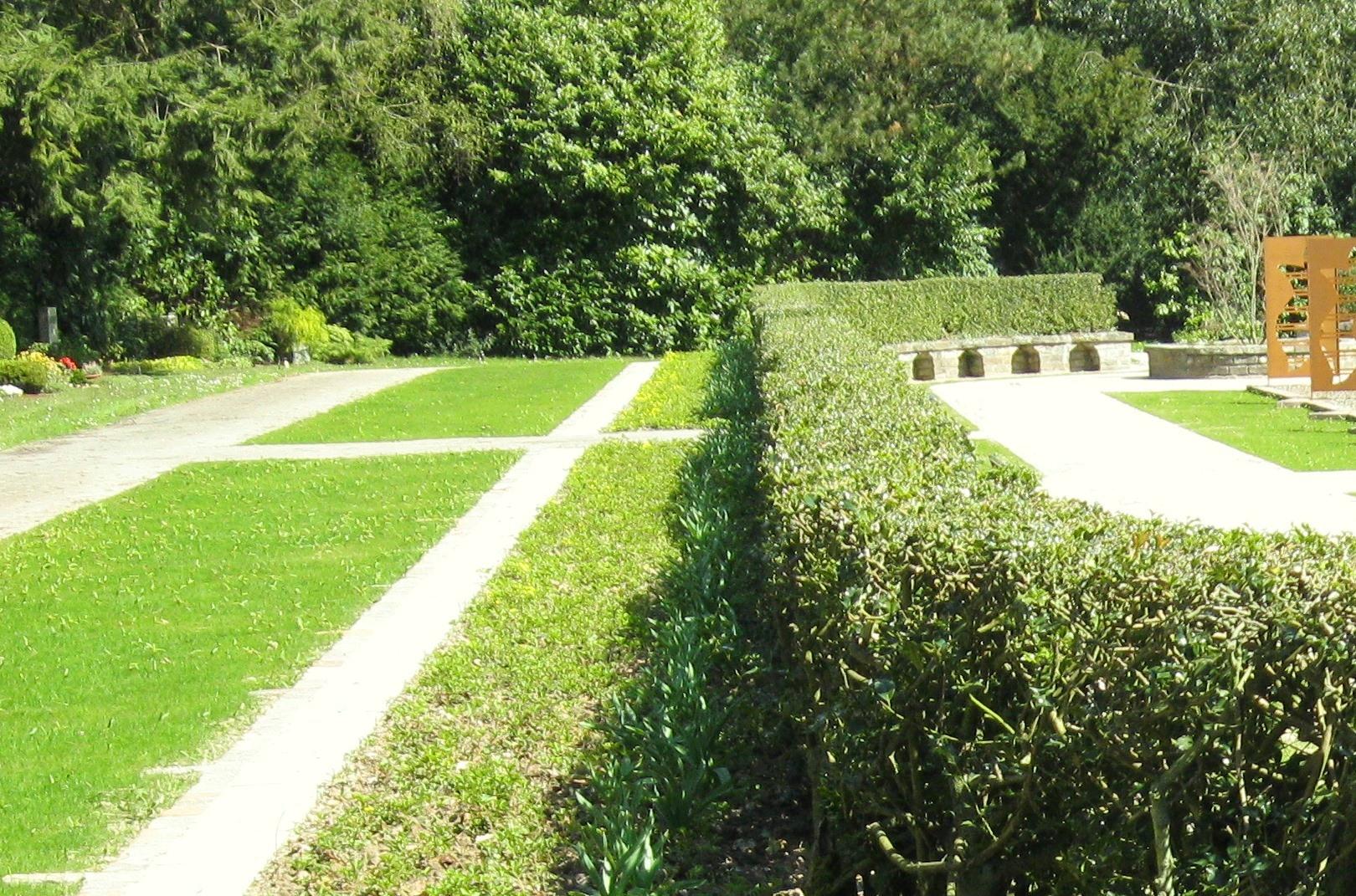 Natursteinmauer, von Hecke umrandet. Daneben Grabfelder für Särge