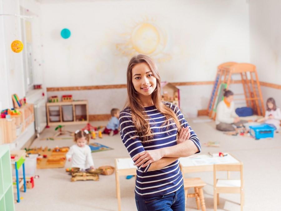 Praktikantin der Kindertagespflege steht in den kindgerecht eingerichteten Räumen, Kleinkinder spielen im Hintergrund. Tagespflegenest, Kita, Kindergarten, Kinderbetreuung, Pflegevater, Pflegemutter.