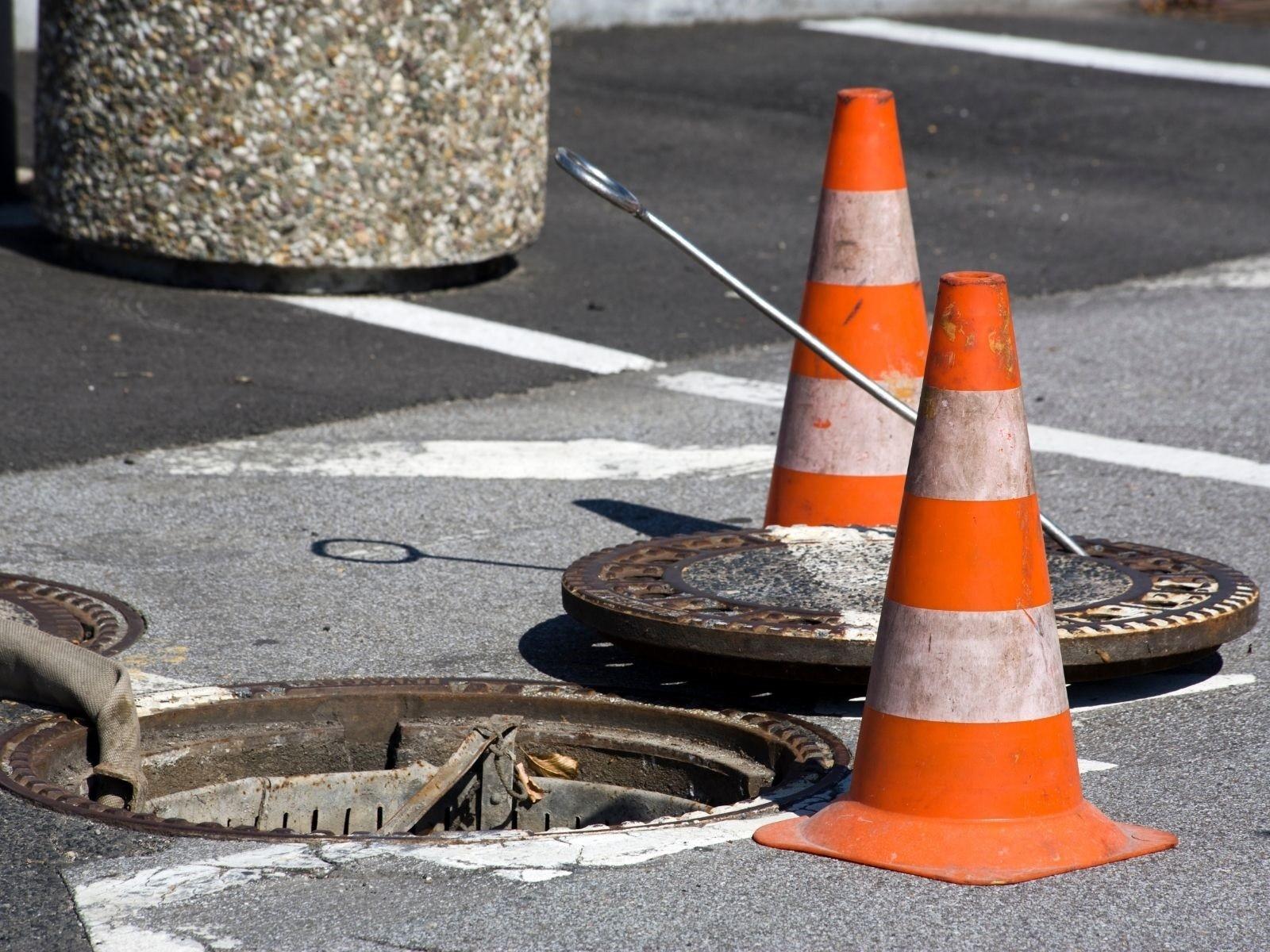 Geöffneter Kanalschacht auf einer Straße, zwei Pylonen schützen die Kanalöffnung.