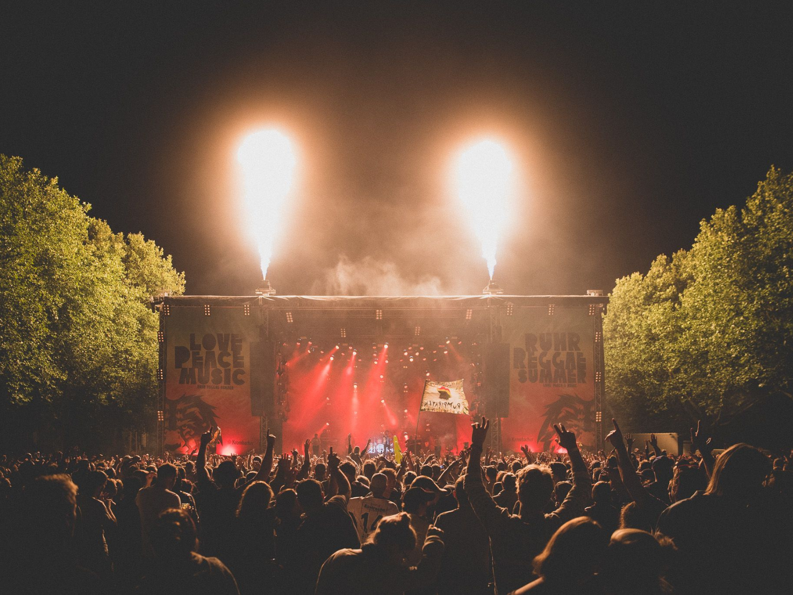 Jubelnde Menge im Dunkeln vor der Bühne beim Ruhr Reggae Summer. Von der Bühne aus schießen zwei Feuerfontänen in den Himmel.