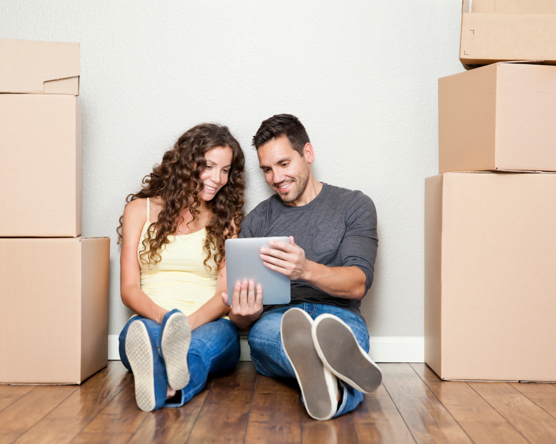 Ein Mann und eine Frau sitzen auf dem Boden zwischen Umzugskartons, schauen auf ein Tablet und unterhalten sich gut gelaunt über das, was sie sehen