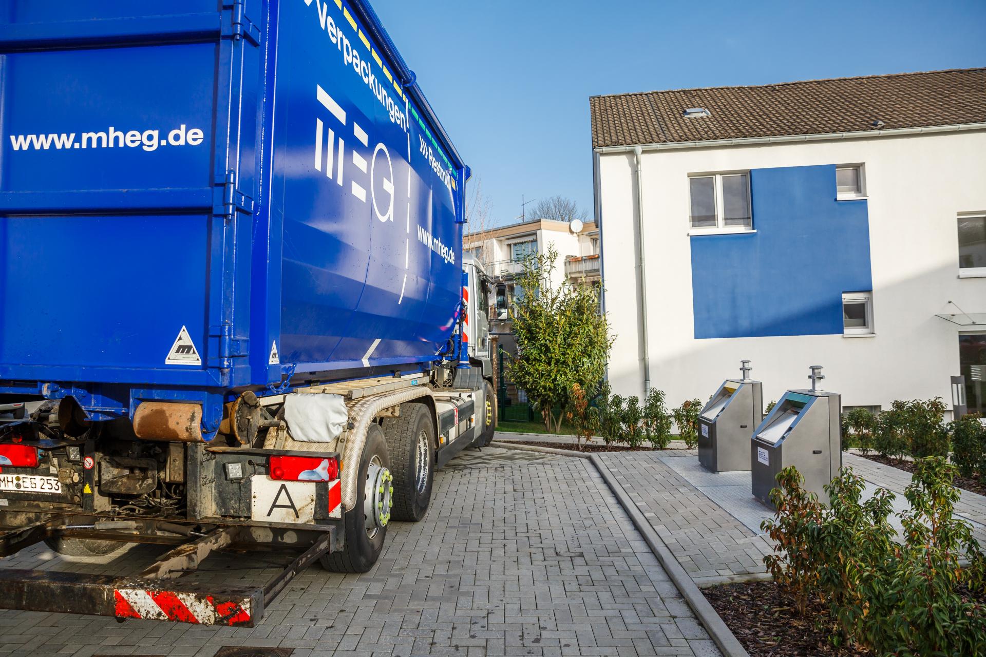 Ein Müllwagen der MEG parkt neben Unterflurbehältern vor einem Wohnhaus.